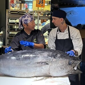 Sakanaya teacher and student prepare to fillet 200-pound tuna laying on the table in front of them.