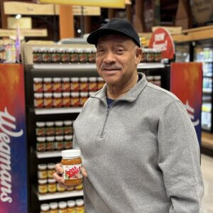Man in grey shirt and black cap holding jar of boss sauce with jars of boss sauce on shelves in the background