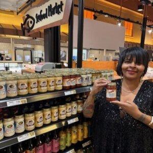 Woman in black top smiling and holding a jar of sauce in front if grocery shelves full of bottled sauces